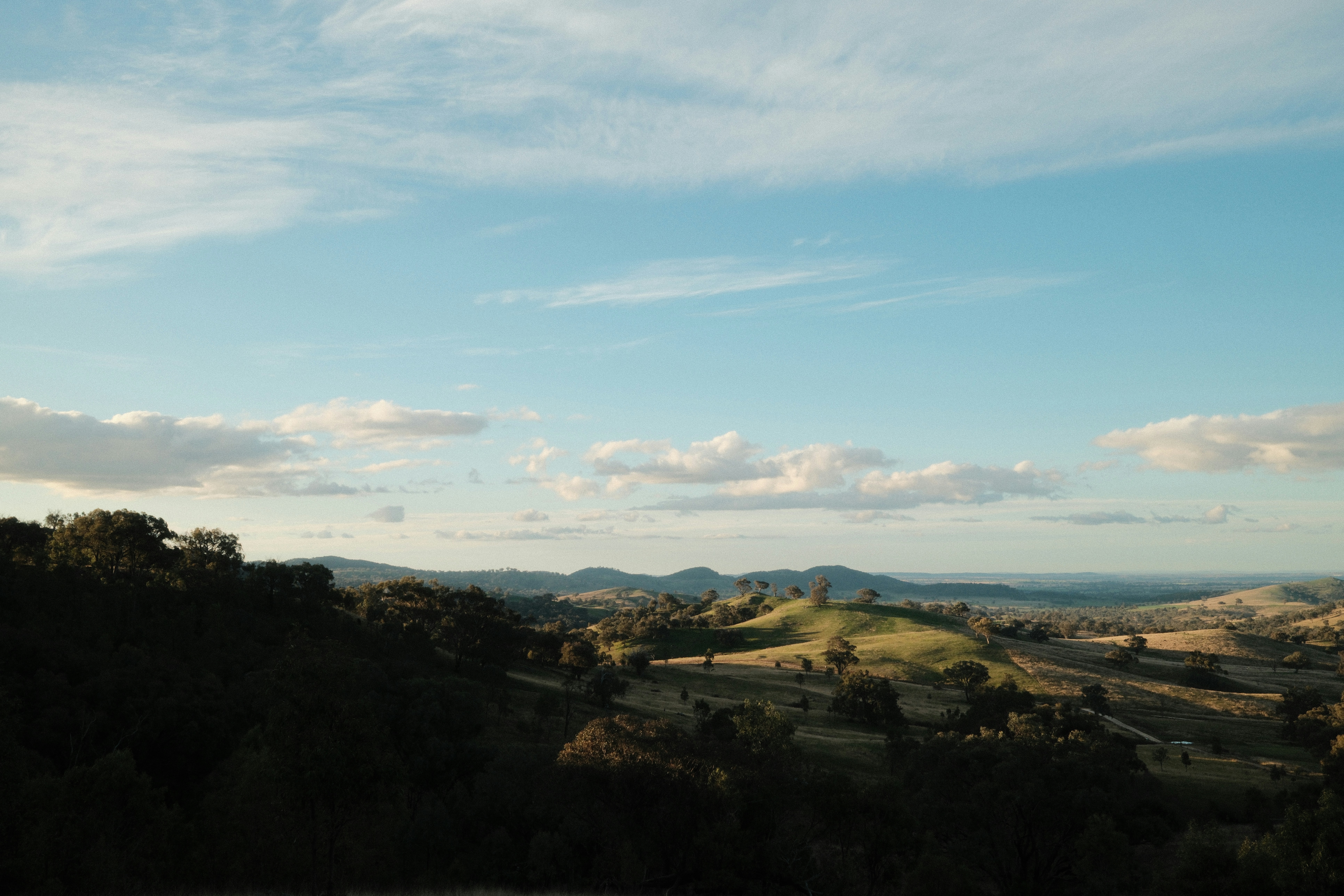 Photo by Mounish Raja taken in Mudgee features rolling green hills with blue sky. A few scattered low clouds hang near the horizon and at the top of the frame. Landscape style.
