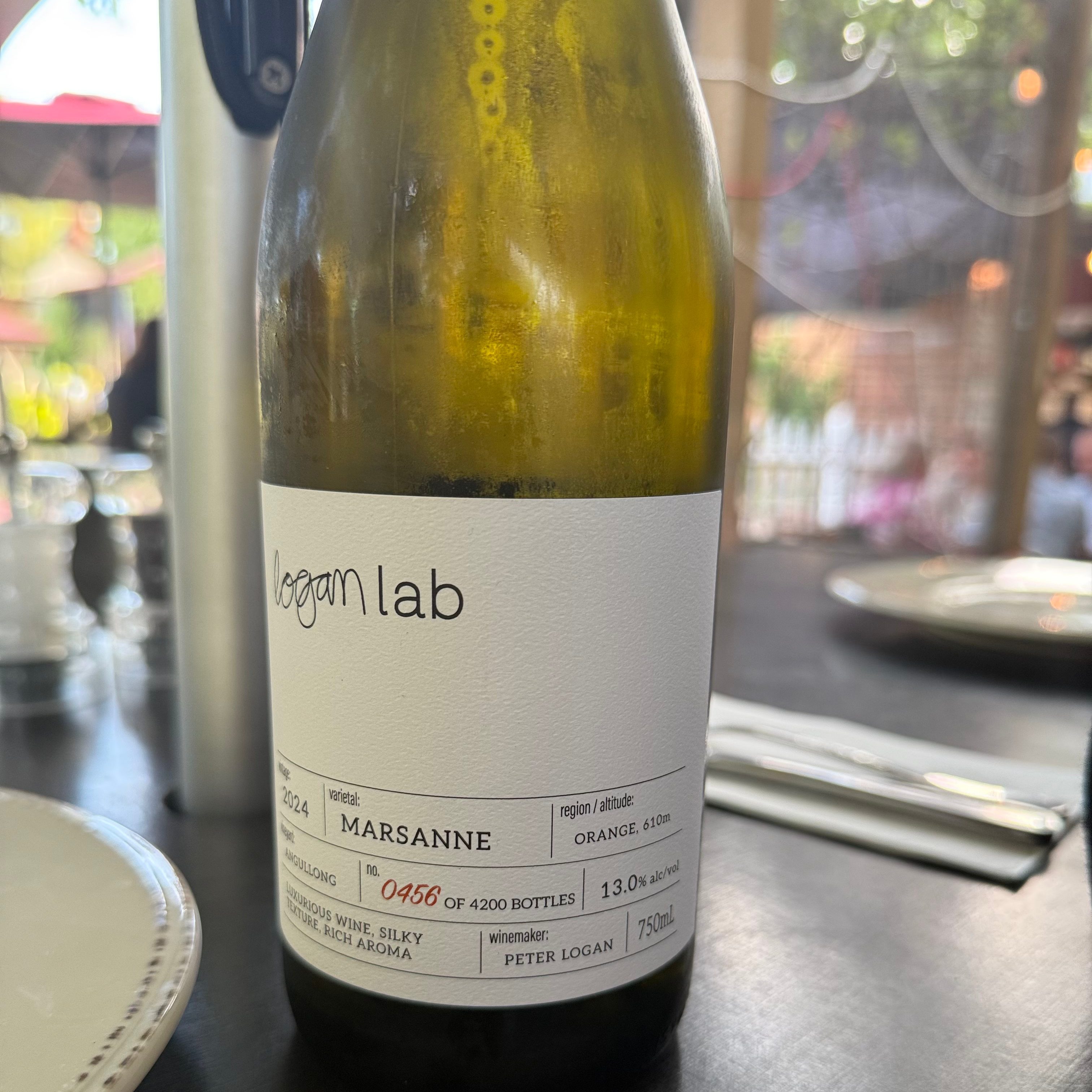 Bottle of Logan Lab wine sitting on a table, visible fairy lights in the background. Wine label says Marsanne bottle 0456 of 4200 bottles.