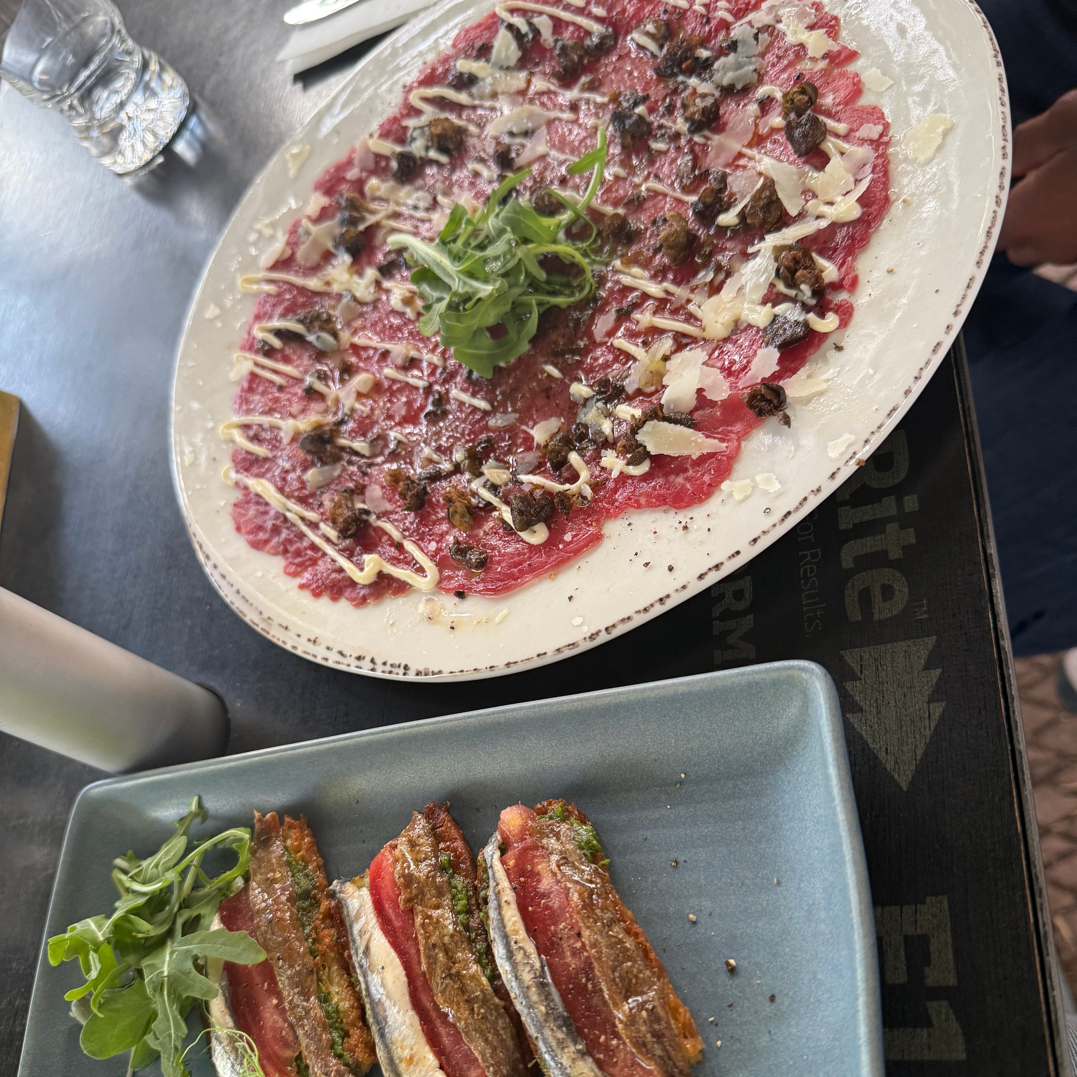 Image of two dinner plats at Food and Farm Dining Mudgee. the plat on the bottom left carries anchovy toast and rocket, and in the top right a plate of beef carpaccio topped with rocket and capers.