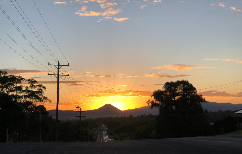 image of the sun rising behind Mount Frome in Mudgee. Taken from the top of Madeira Road at Flirtation Hill Lookout. Powerlines and trees are visible on the left. A tree silhouetted on the right.