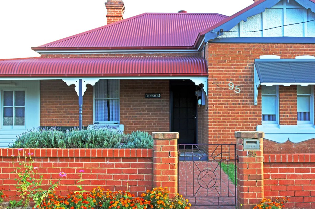 Image shows front facade of Merala house, red brick and red tin roof. Brick fence.