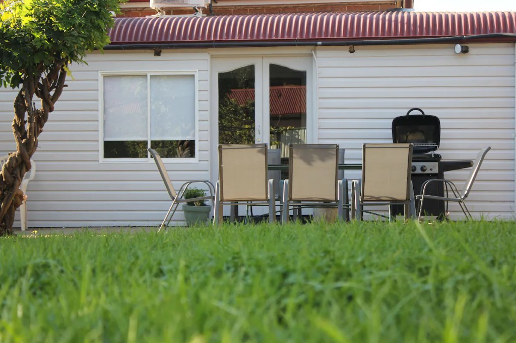 Image shows green grass in backyard and table and chairs, rear of building is visible in the background.