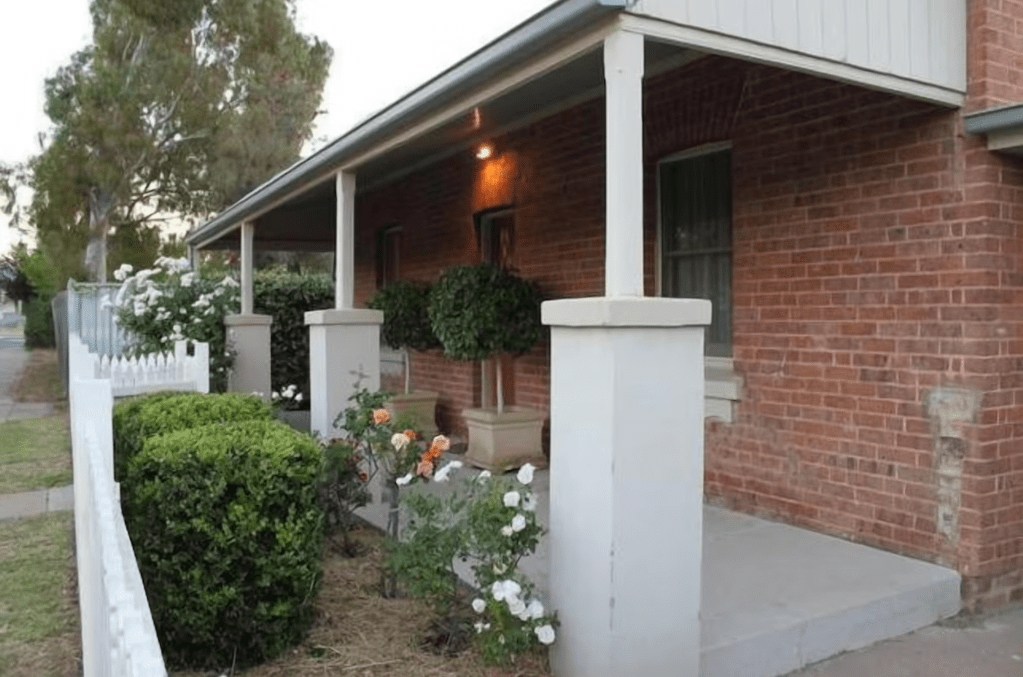 Side view of the front verandah of Douro Cottage, Central Mudgee. Rose garden and cement pillars contrast green hedges and white picket fence slightly visible. 